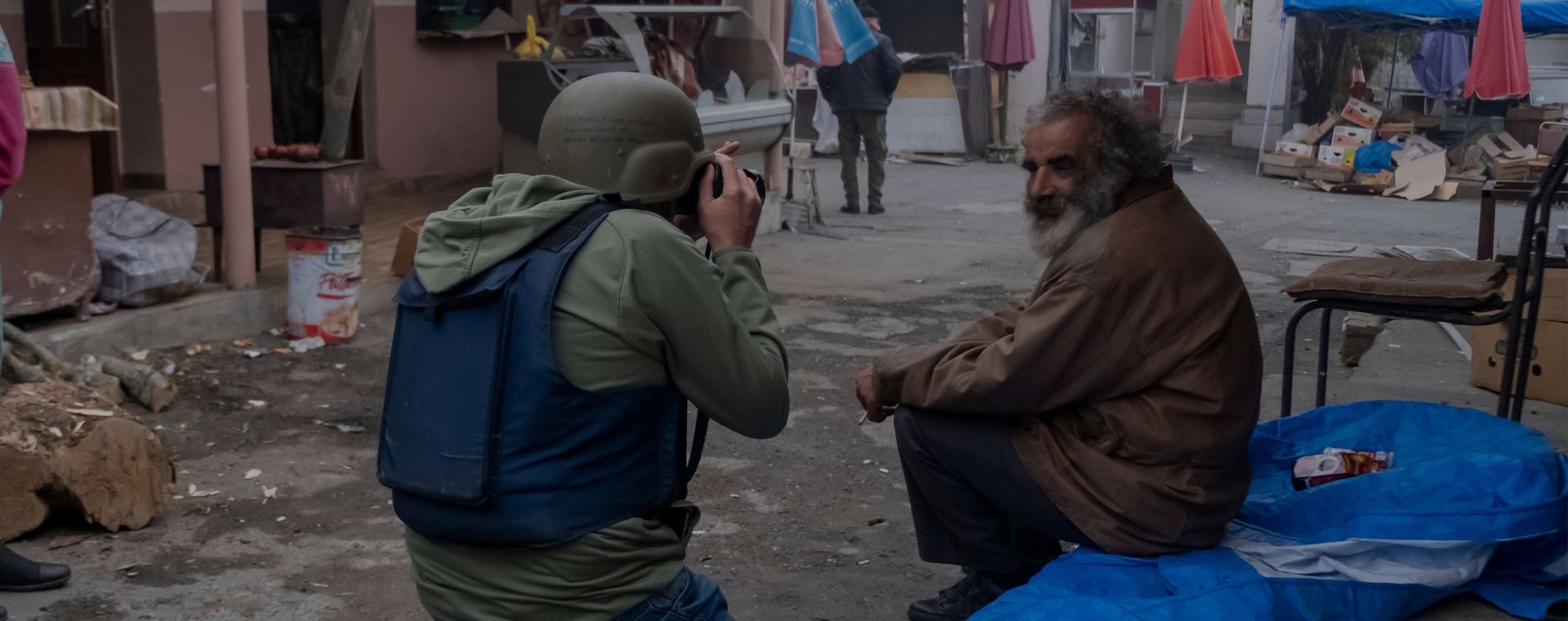 A photographer wearing protective flak jacket and helmet photographing an elderly local man during a military conflict between Armenian and Azerbaijani forces at the central market in Stepanakert